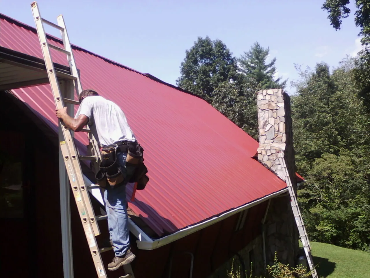 Roofer on a ladder beside a red metal roof and stone chimney — Soffit & Fascia Repair by Roofing Brothers in Weiser